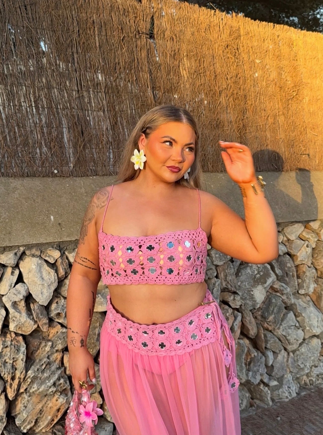A woman wearing a pink two-piece crochet sarong set with silver mirror embellishments, consisting of a cropped bandeau top and a matching sheer skirt, posing outdoors against a stone wall and thatched wall background.